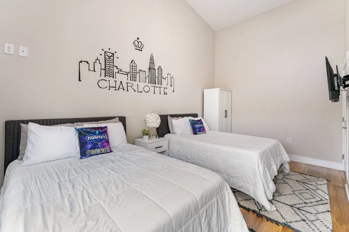 A stylish loft bedroom with two comfortable beds and decorative pillows. Above the beds hangs art depicting the city skyline, and a nightstand with a lamp sits between them, further emphasizing the urban location and contemporary design.