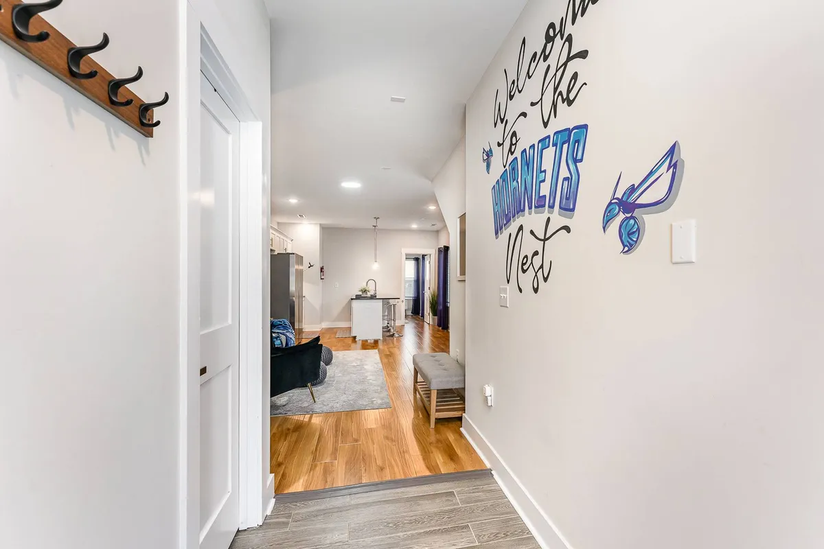 The image presents a hallway with 'Welcome to the Hornets Nest' wall art, leading into the kitchen. The design celebrates local sports culture, and the energetic atmosphere makes it clear this is a place for fans and social gatherings.