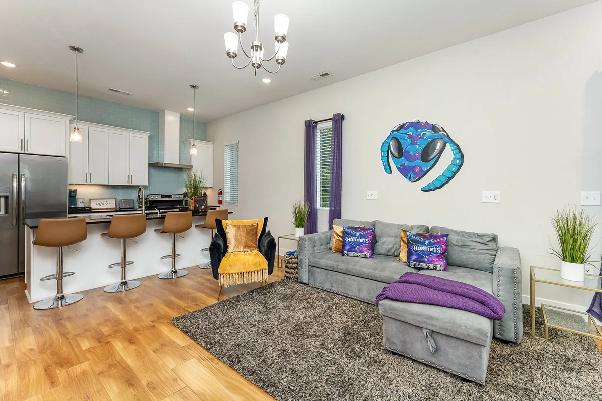 A stylish, comfortable living space featuring a plush sofa with a purple throw, a sleek kitchen island with stools, and colorful wall art. The layout highlights an open, social atmosphere, ideal for both relaxing and entertaining.