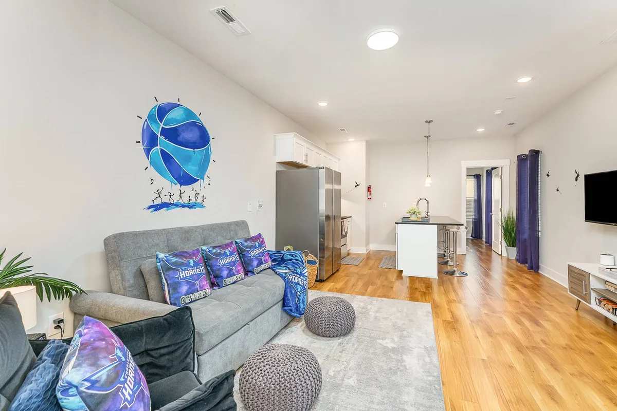 This interior image features a colorful basketball mural that sets a playful tone, a comfortable gray couch with decorative pillows, and a modern kitchen island in the background. The open space is inviting, with hints of social activity and entertainment throughout the decor.