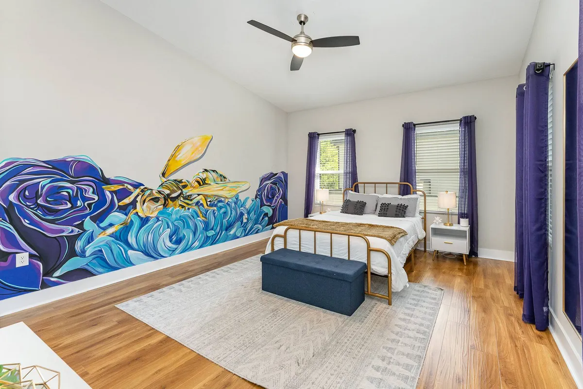 A vibrant bedroom with a golden bed frame set against a colorful mural of roses. A blue storage ottoman sits at the foot of the bed, and the room exudes a warm, artistic, and serene atmosphere—ideal for unwinding in privacy after a day of activity.