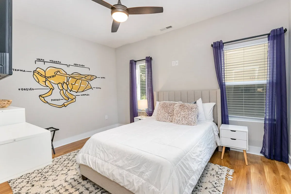 A cozy bedroom featuring a neatly made bed with plush pillows, a whimsical lobster illustration on the wall, and a window with soft purple curtains letting in natural light. The room feels restful and well-appointed, perfect for a peaceful night's sleep or quiet relaxation.