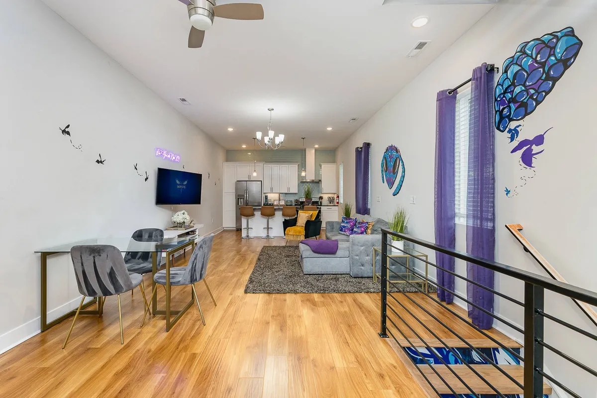 A bright, open living space featuring a stylish sofa, eye-catching wall art, a visible dining area, a kitchen, and large windows that flood the space with natural light. The room feels lively and welcoming, ideal for group gatherings and meals.