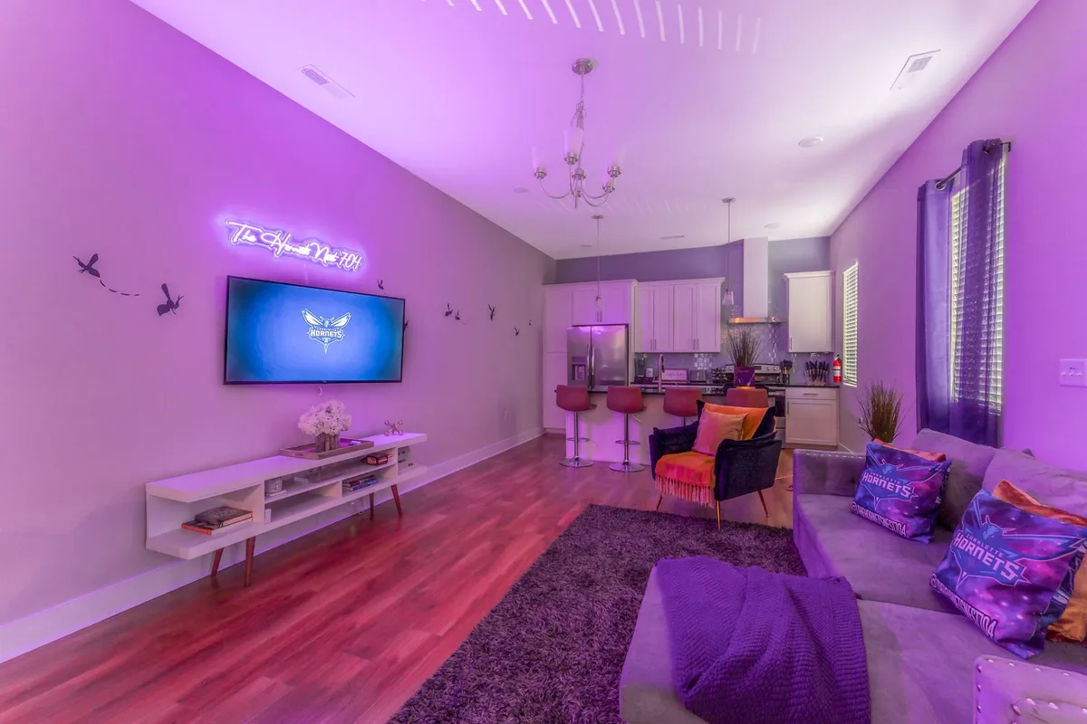 A vibrant living area with a plush, comfortable sofa, colorful accent chairs, a striking neon sign on the wall, and a stylish rug. The television is positioned for group viewing, and the seating arrangement encourages lively conversation and gatherings.