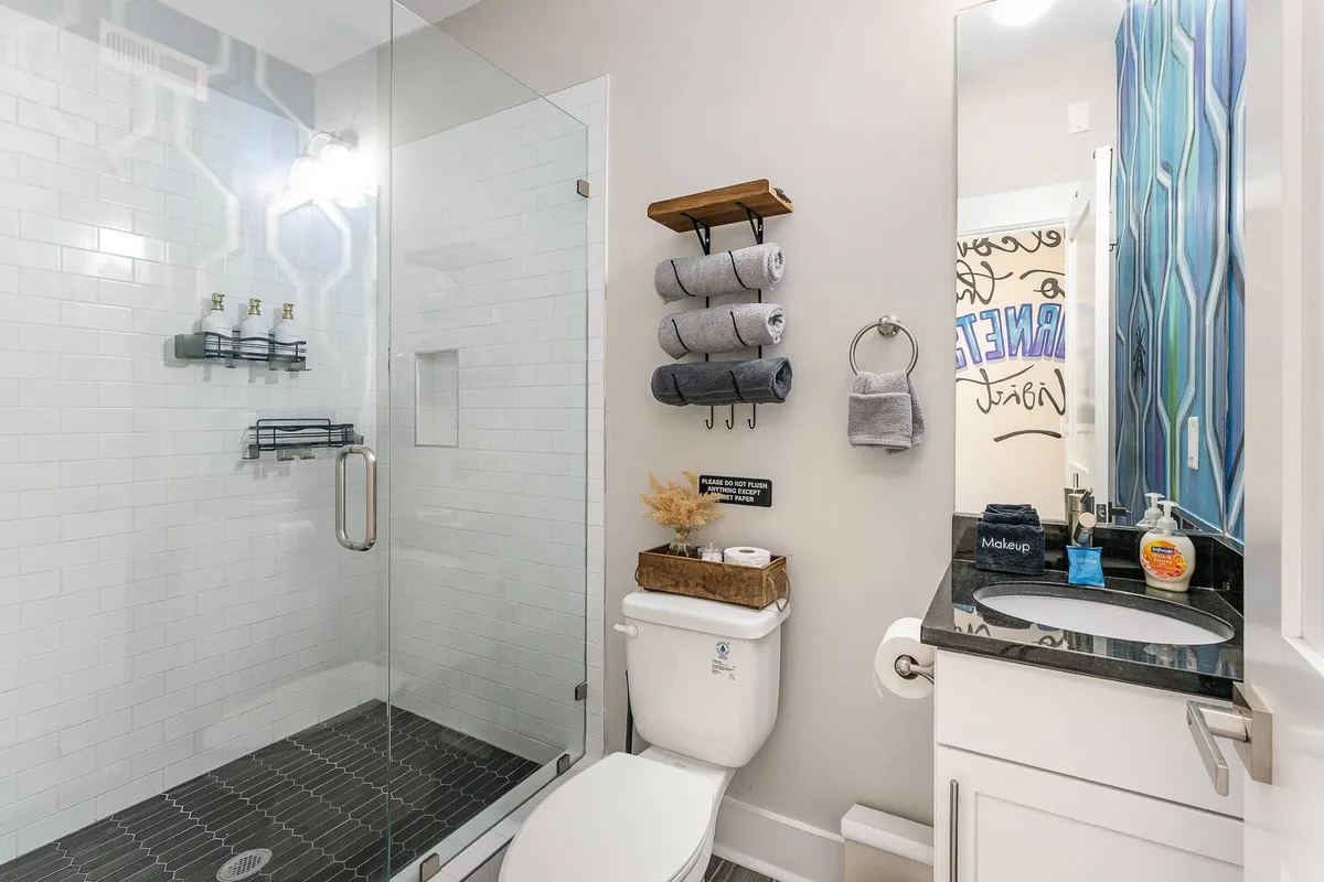 A stylish, contemporary bathroom with a glass shower enclosure, dark patterned floor tiles, a floating wooden shelf stacked with towels, and a vanity with a mirror. The space feels modern, clean, and spa-like, ideal for rejuvenation.