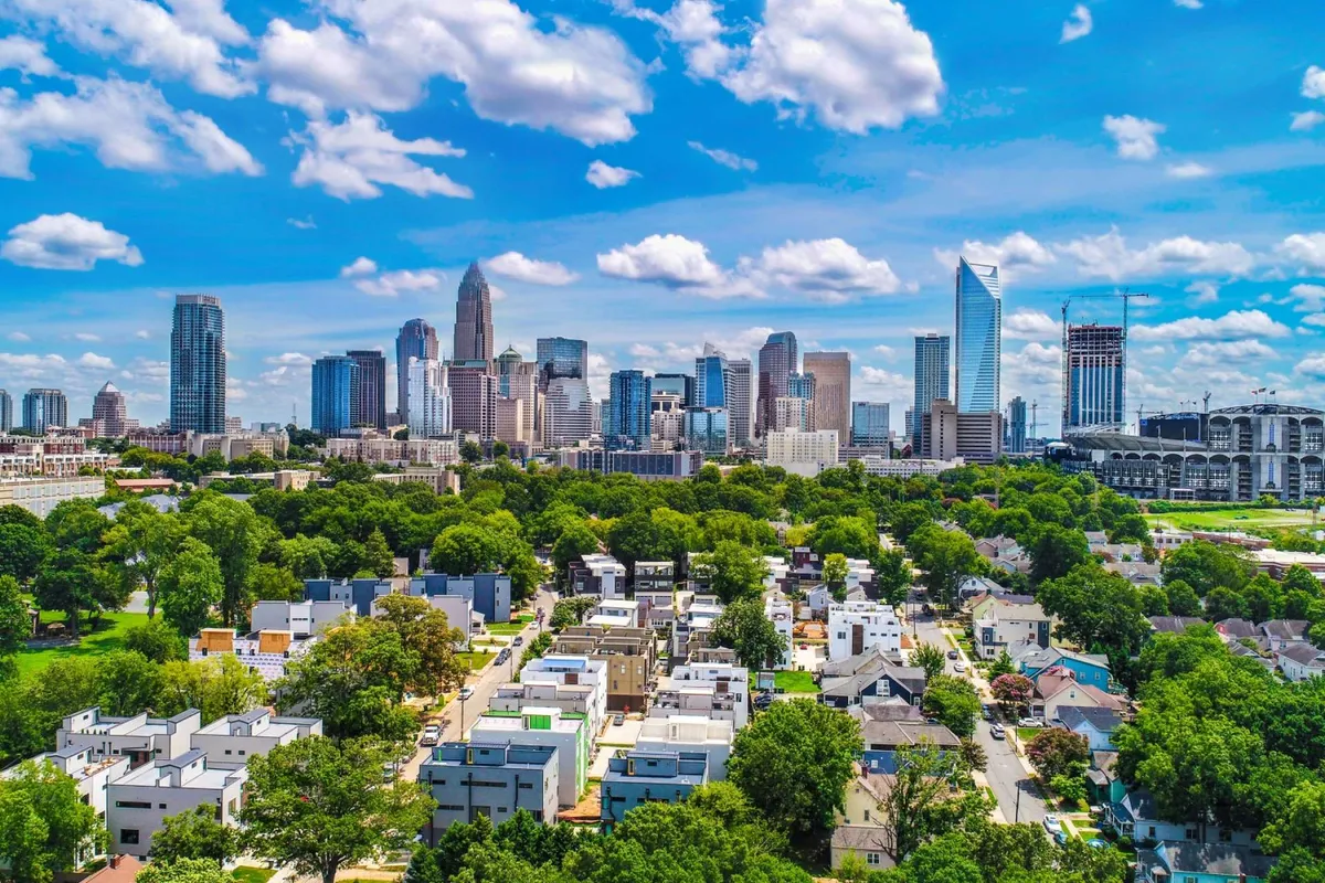 A sweeping daytime view of the Charlotte city skyline, green parks, and nearby residential areas, as seen from an elevated perspective. The energetic cityscape highlights the vibrancy and proximity of Uptown Charlotte.