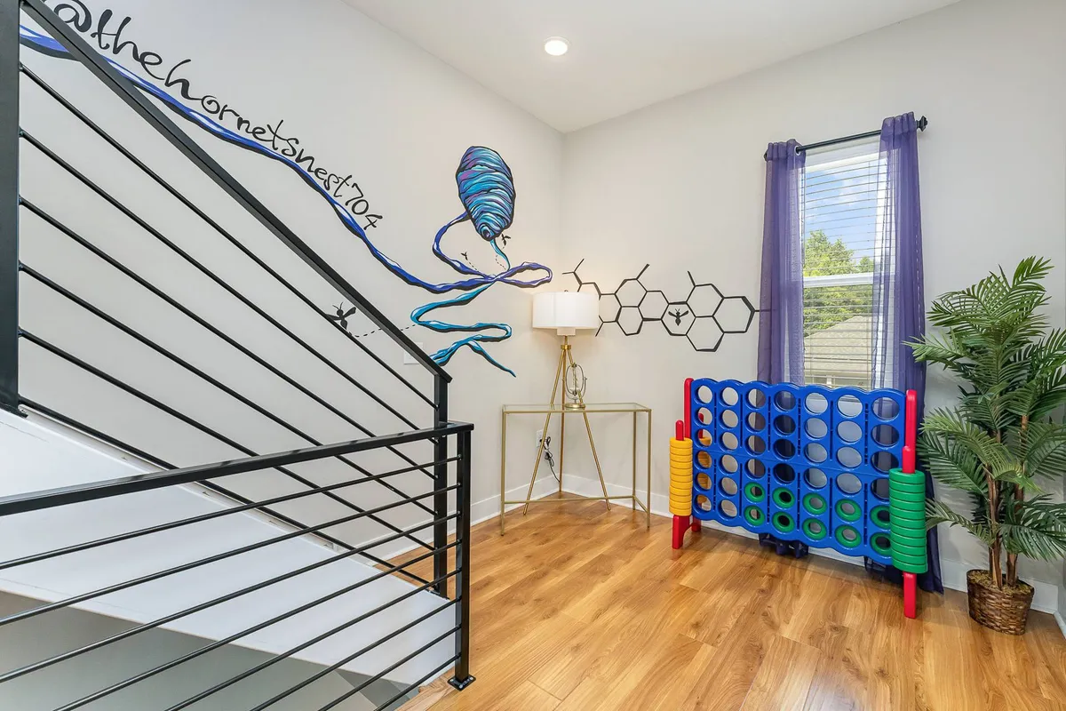 A playful interior scene featuring a staircase railing, a giant Connect 4 game set up for play, a decorative lamp, and colorful wall mural. The area is energetic and clearly provides space for interactive entertainment and socializing.