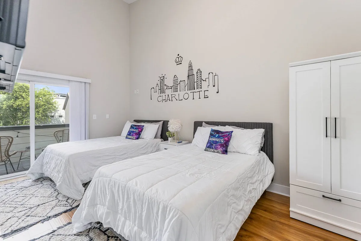 A bright, airy room with two beds made up with decorative pillows, art featuring a city skyline on the wall, and sunny morning light flooding in. The space is welcoming and perfect for children, cousins, or friends to share, reflecting a playful yet restful environment.