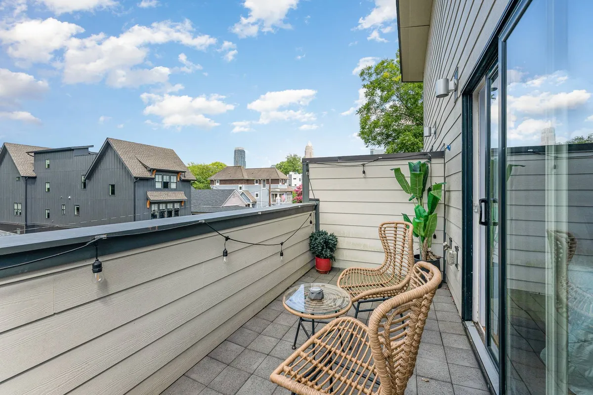 A cozy private balcony furnished with wicker chairs and a small table, accented by a potted plant. The balcony offers a sweeping city view, with the Uptown Charlotte skyline visible in the background, and the warm hues of sunset casting a tranquil glow over the space.